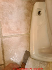 Cutting under the toilet bottom edge to separate it from the adhesive sealant at the floor (C) Daniel Friedman at InspectApedia.com